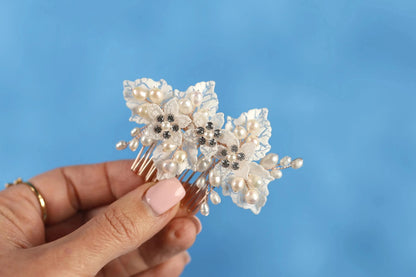 A hand holding a decorative hair accessory with white flowers and pearls against a blue background.