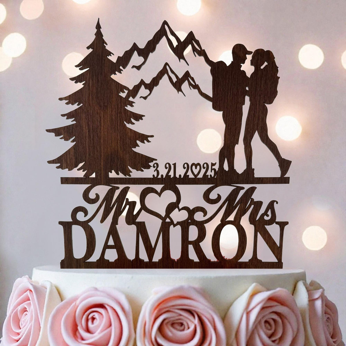 A wooden wedding cake topper featuring a silhouette of a couple embracing, with mountains in the background and the names 'Mr. and Mrs. DAMRON' written below.