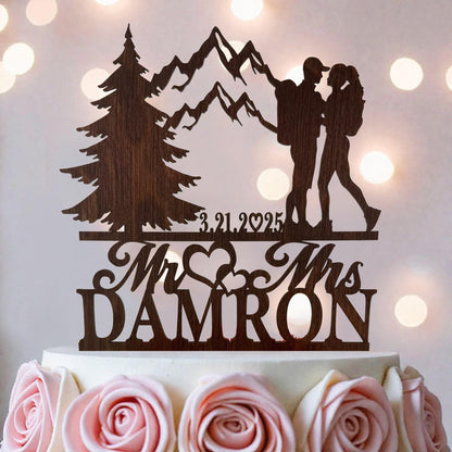 A wooden wedding cake topper featuring a silhouette of a couple embracing, with mountains in the background and the names 'Mr. and Mrs. DAMRON' written below.
