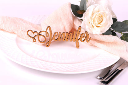 A wooden nameplate with the name "Jennifer" on it, placed on a white plate with a pink napkin and a white rose.