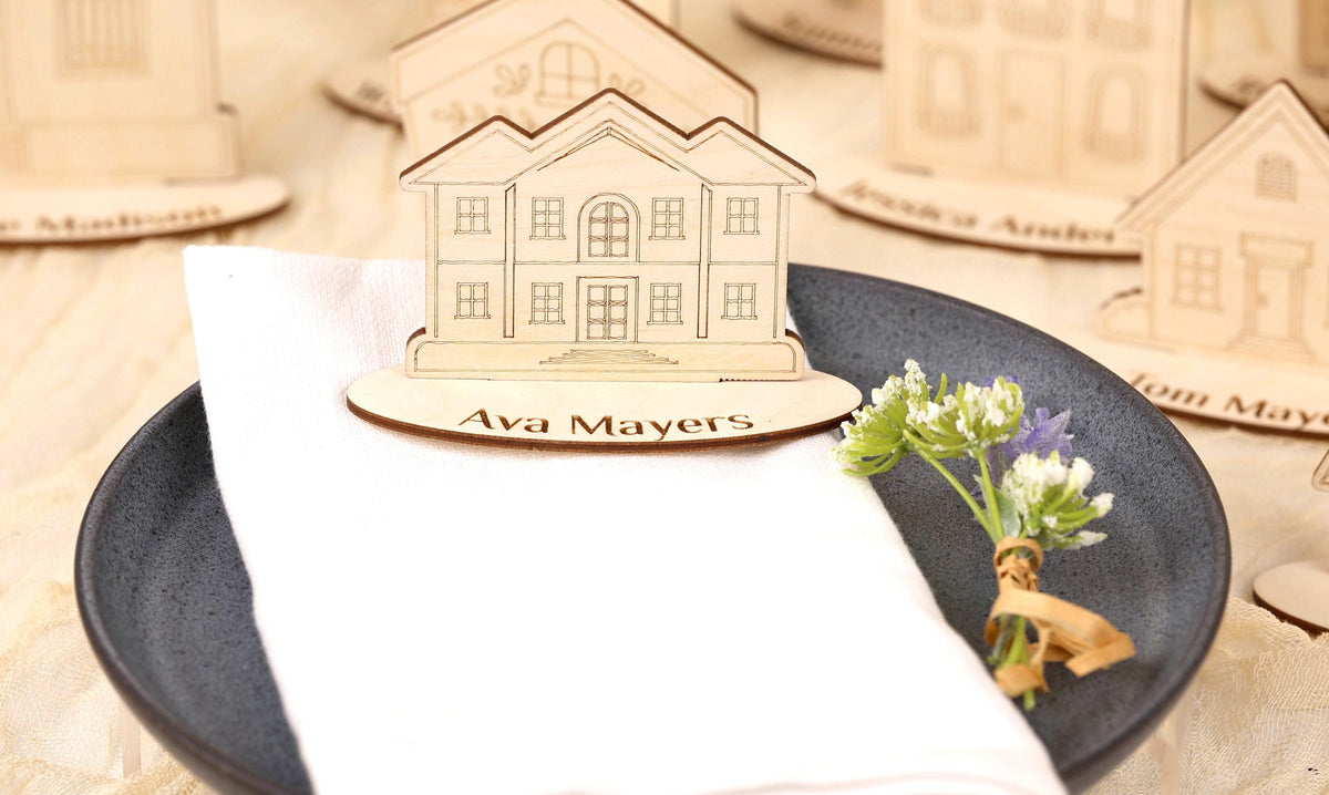 A wooden house-shaped nameplate with the name "Ava Mayers" is placed on a white napkin, accompanied by a small bouquet of flowers.
