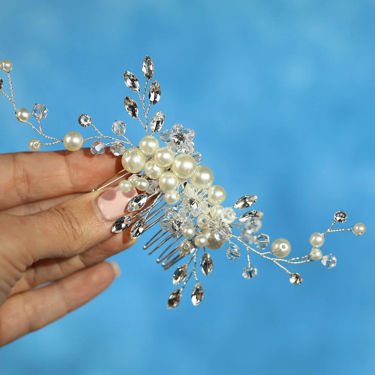 A hand holding a decorative hair accessory with pearls and crystals against a blue background.