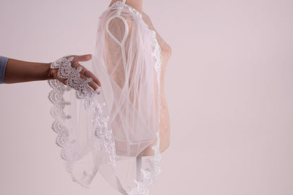 A hand holding a delicate white lace veil with a scalloped edge, against a plain white background.