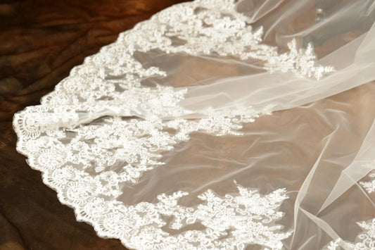 A close-up view of a delicate white lace veil with intricate floral embroidery, resting on a wooden surface.