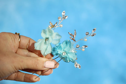 A hand holding two delicate flower-like objects with sparkling crystal or gemstone accents against a blue background.