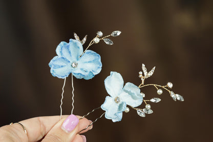 A hand holding two delicate flower-shaped hair clips with blue petals and sparkling crystal embellishments.