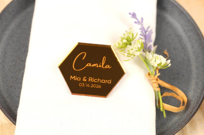 a table setting with a white napkin, a black plate, and a small floral arrangement. There is a hexagonal nameplate with the name 'Camila' and the date '03.14.2026' on it, placed on the napkin.