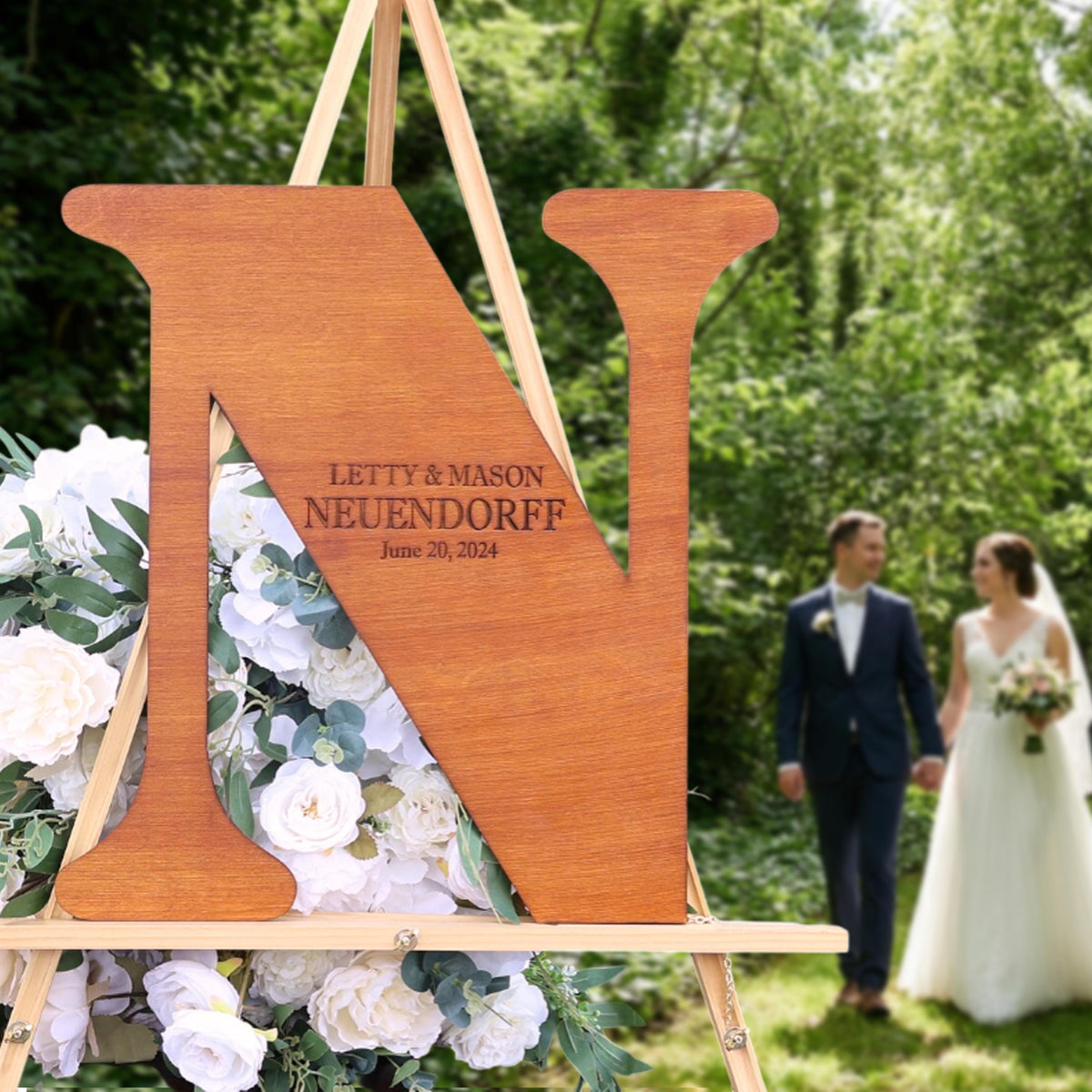 A wooden sign with the initials "N" and the names "LETTY & MASON NEUENDORFF" is displayed in a garden setting, with a couple walking in the background.