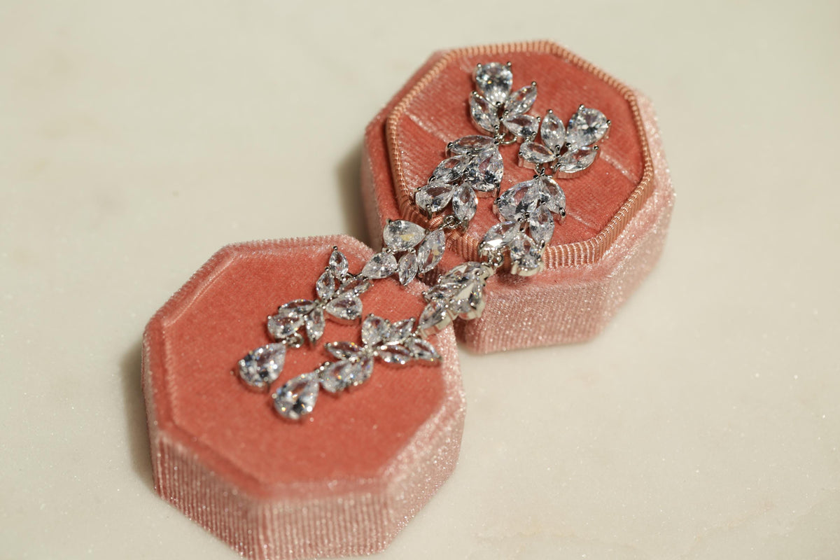 A pair of diamond earrings in a pink velvet box, with the earrings hanging from the box.