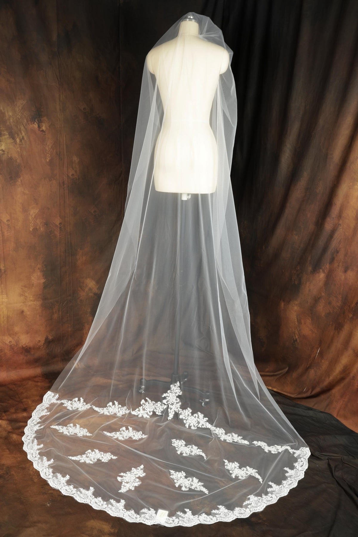 A white wedding veil with a lace trim is displayed on a mannequin against a brown background.