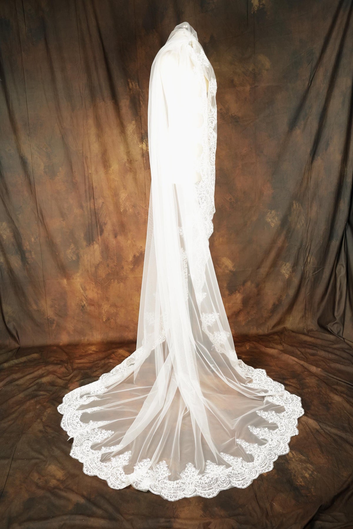 A white wedding veil with a long train and a lace trim is displayed against a brown background.