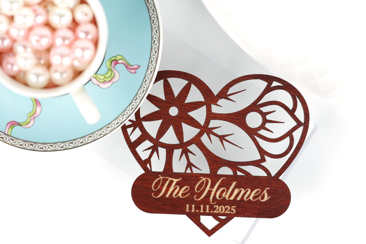 a wooden heart-shaped card with the name "The Holmes" and the date "11.11.2025" printed on it, placed next to a teacup filled with pink pearls.