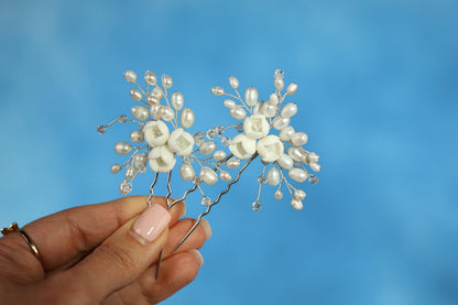 A hand holding a pair of decorative hair clips with white pearls and silver beads against a blue background.