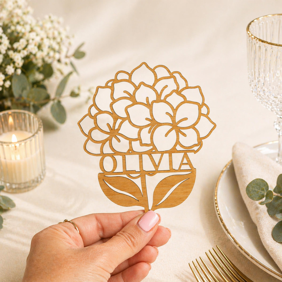A hand holding a wooden cutout with the name "Olivia" on it, surrounded by floral designs and a candle on a table.