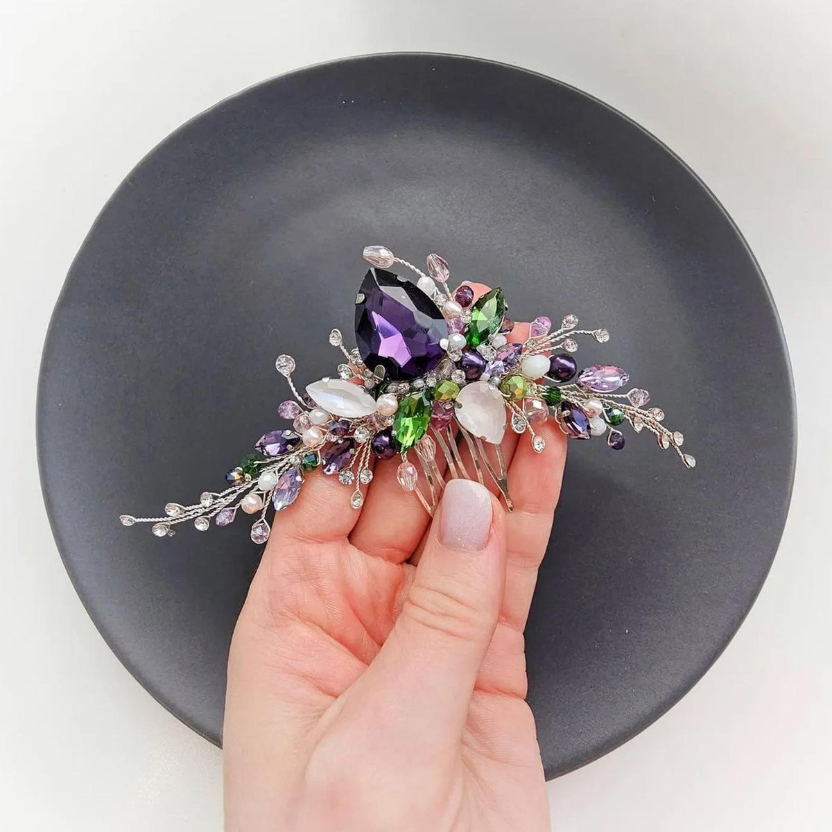 A hand holding a decorative hair accessory with a variety of colorful gemstones and pearls.