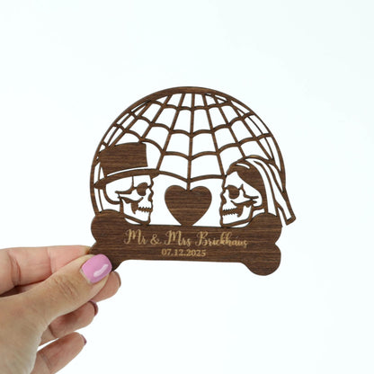 A hand holding a wooden cutout with a design featuring two skull faces and a heart shape, along with the text "Mr & Mrs Brickhaus 07.12.2025".