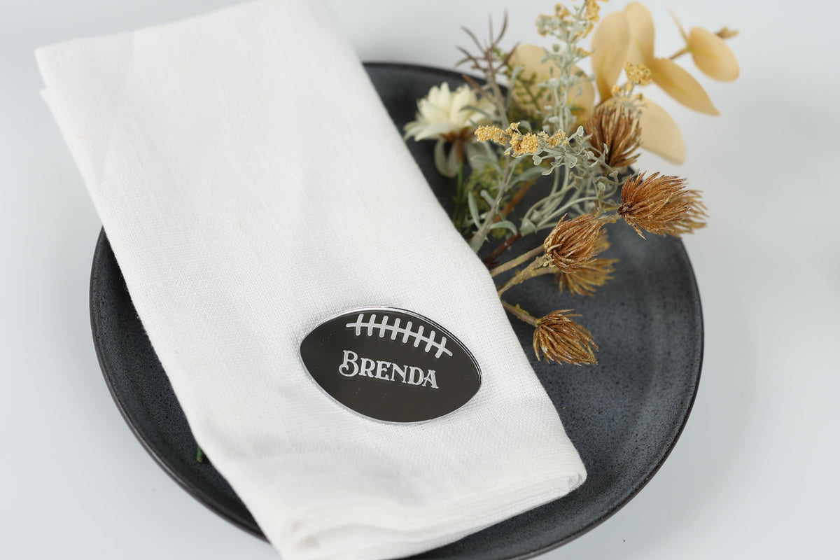 A white napkin with a black oval-shaped name tag that says "BENDA" is placed on a black plate, with some dried flowers and yellow flowers nearby.