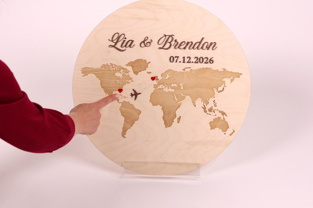 A person is pointing to a wooden sign with a world map and the names 'Lia & Brandon' and the date '07.12.2026' on it.