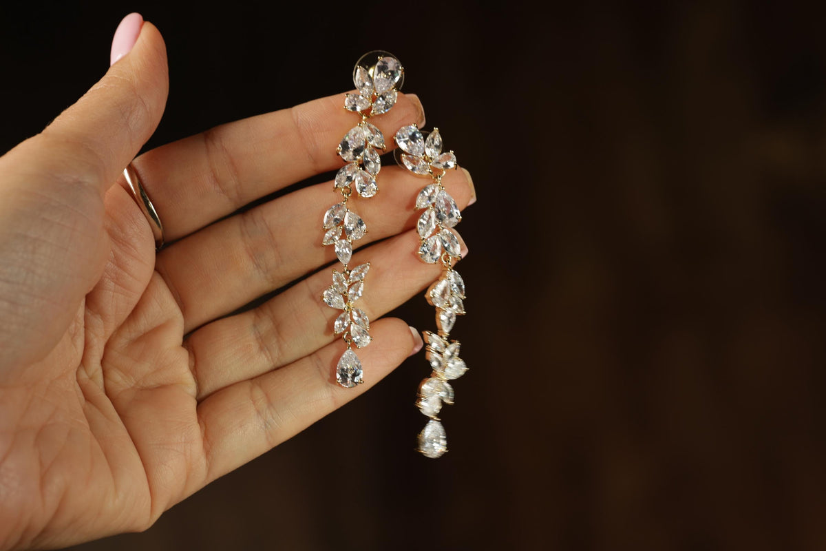 A hand holding a pair of elegant gold earrings with numerous small, clear gemstones.