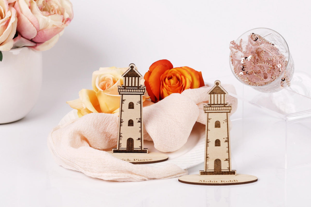 two wooden lighthouse-shaped objects placed on a white surface, surrounded by a vase of pink roses and a glass container with pink glitter.