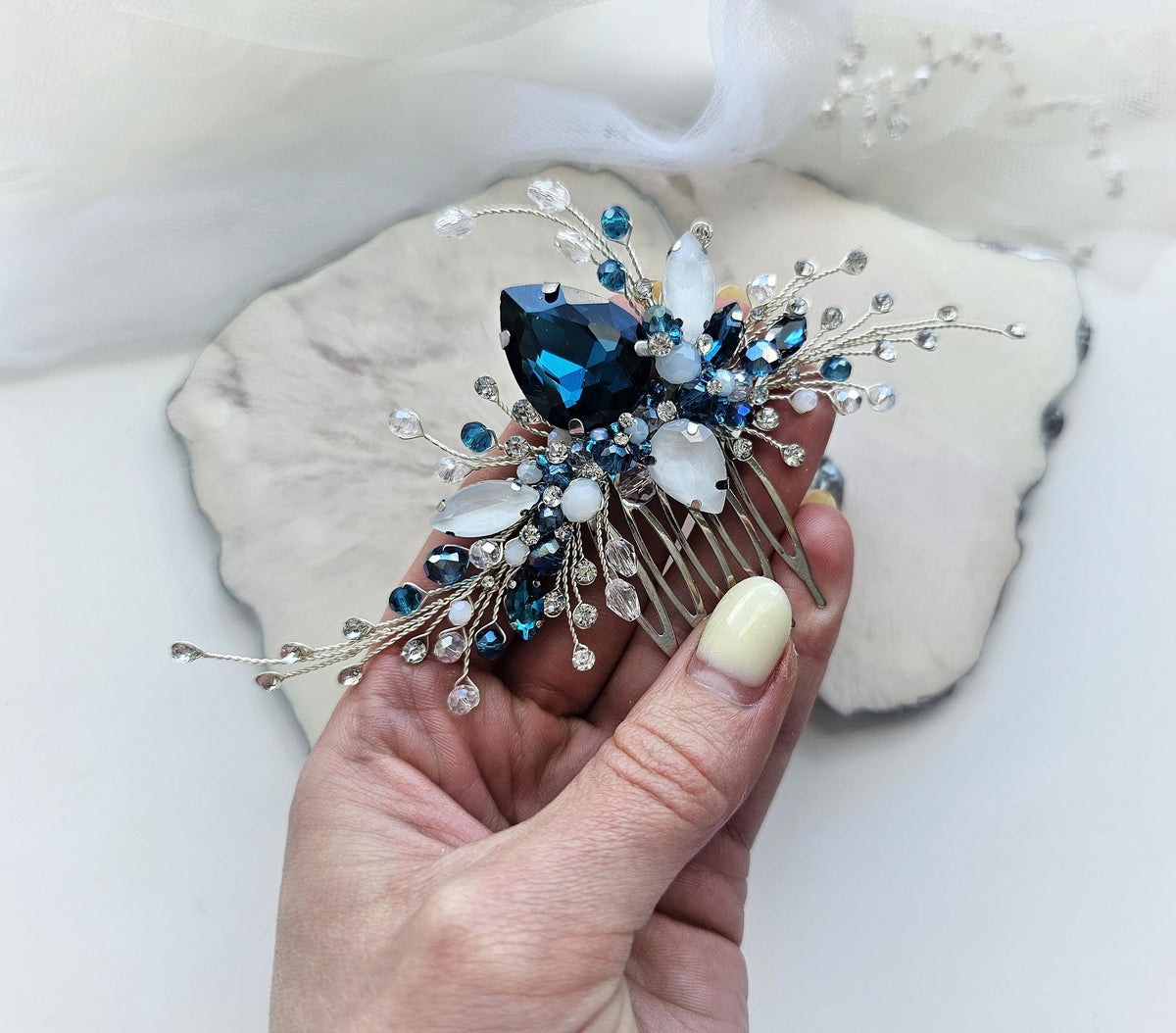 A hand holding a decorative hair accessory with blue and white gemstones and silver metal.