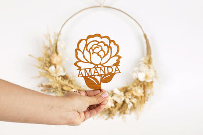 A hand holding a wooden rose-shaped cake topper with the name "AMANDA" written on it.