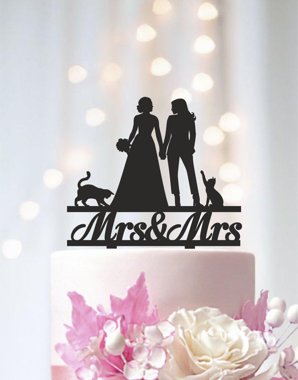 A silhouette of a bride and groom holding hands, with a cat and a flower on a cake topper, against a blurred background with lights.