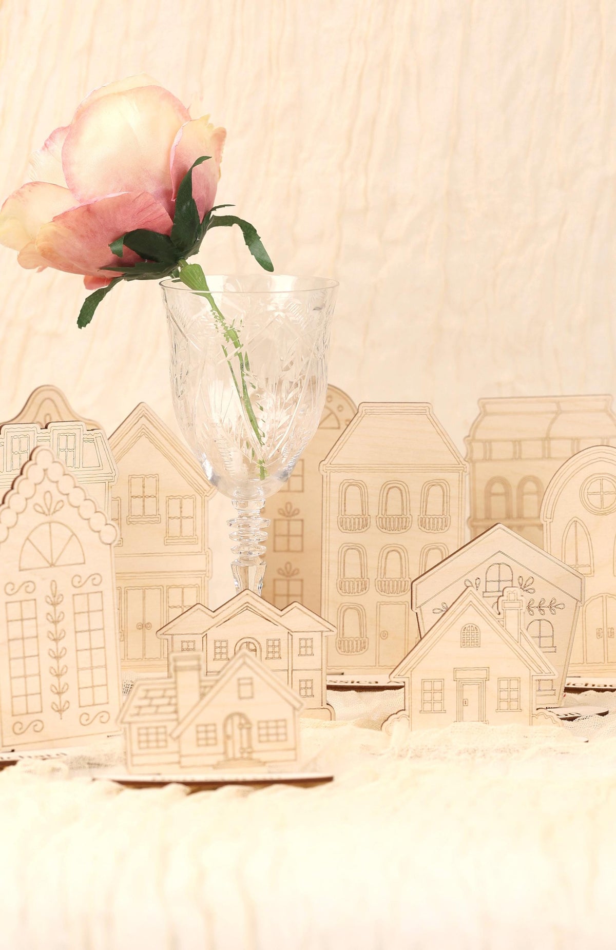 A single pink rose in a clear glass vase is placed in front of a collection of wooden cutouts depicting houses, creating a charming and decorative scene.