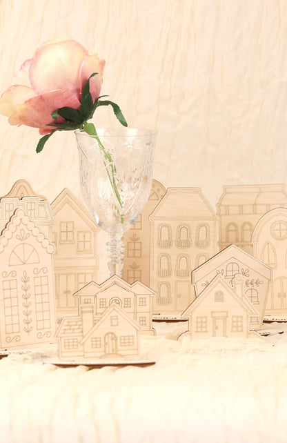 A single pink rose in a clear glass vase is placed in front of a collection of wooden cutouts depicting houses, creating a charming and decorative scene.