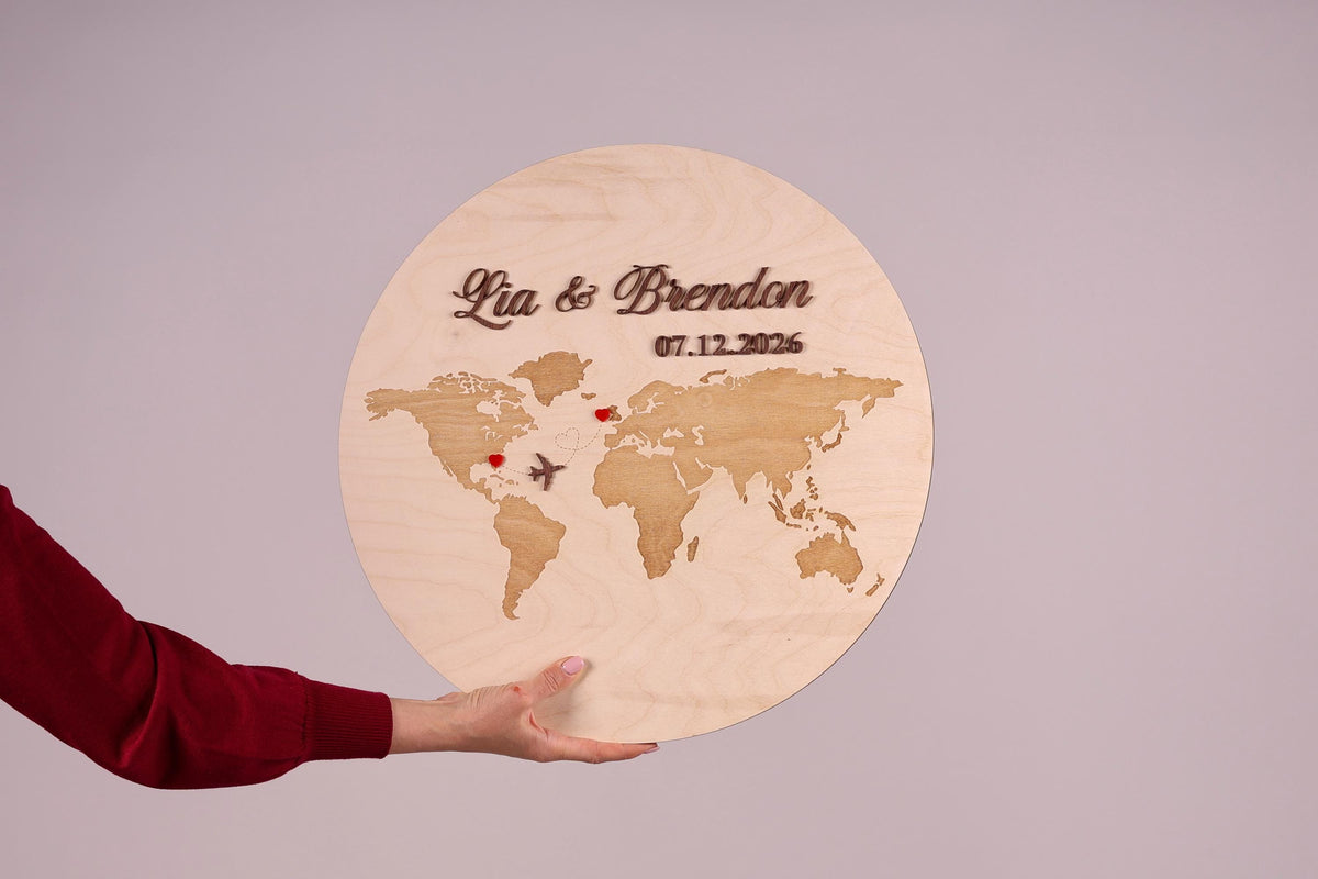 A person's hand holding a wooden sign with a world map and the names 'Lia & Brendan' written on it.