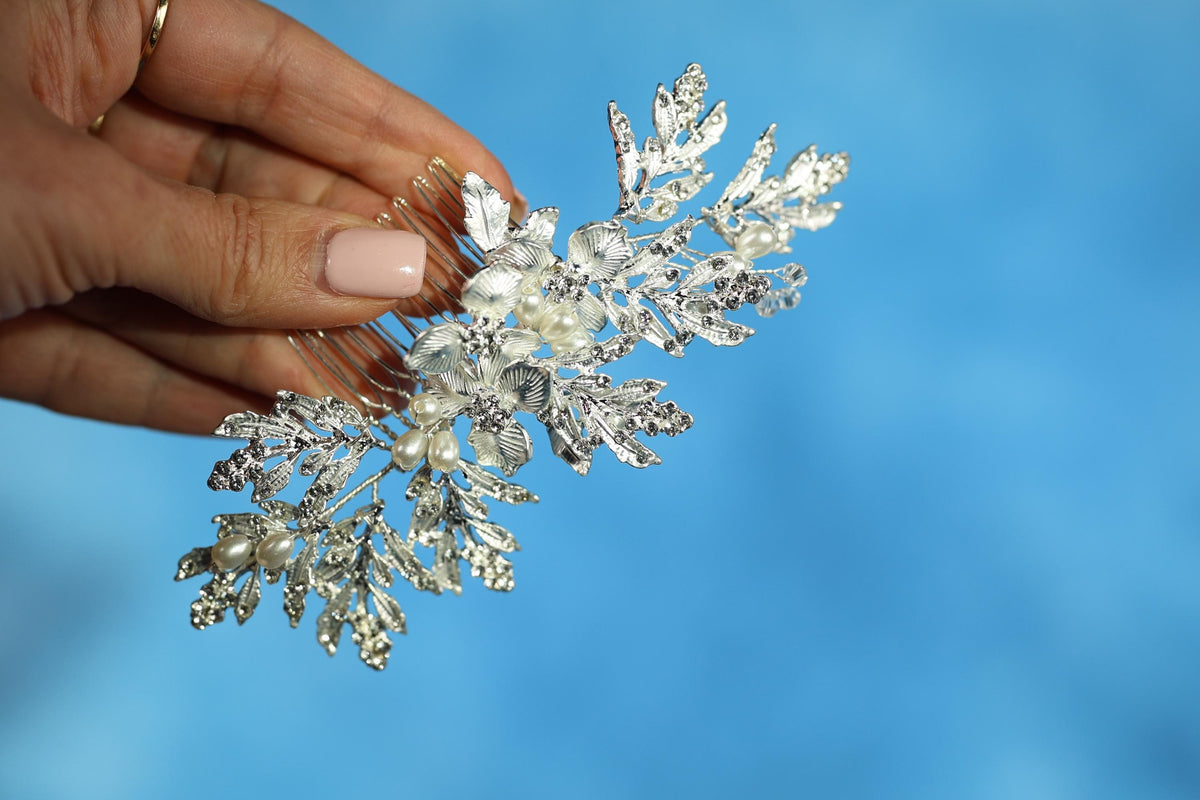 A hand holding a decorative silver hair accessory with leaves and flowers against a blue background.