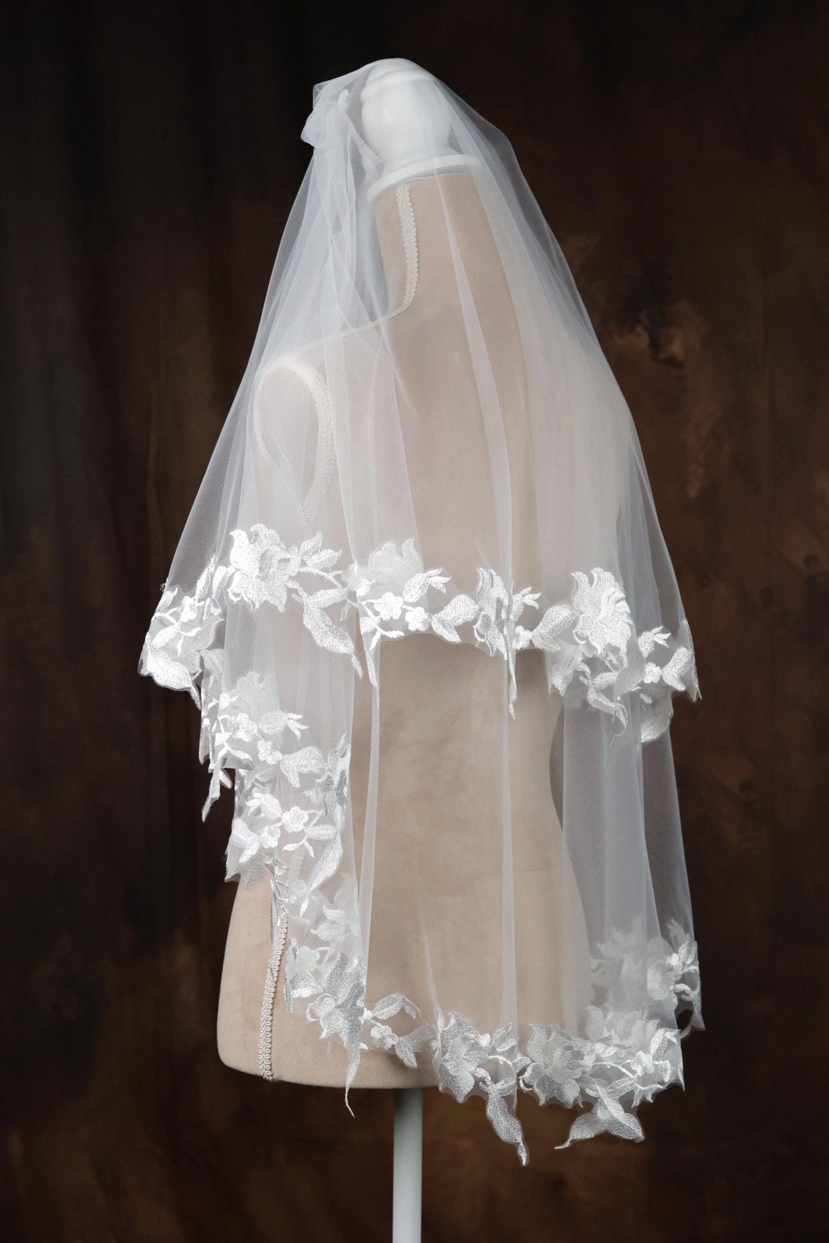 A white wedding veil with a floral lace trim is displayed on a mannequin against a dark background.