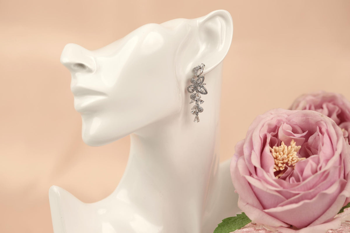 A white mannequin head wearing a pair of dangling earrings with a floral design, placed next to a pink rose.