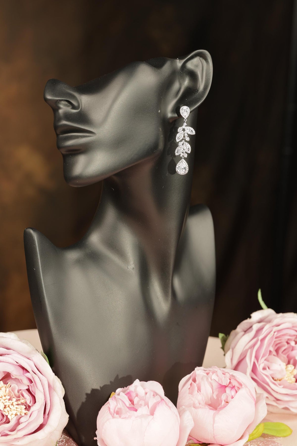 A black mannequin head with a pair of earrings on it, placed on a surface with pink peonies.