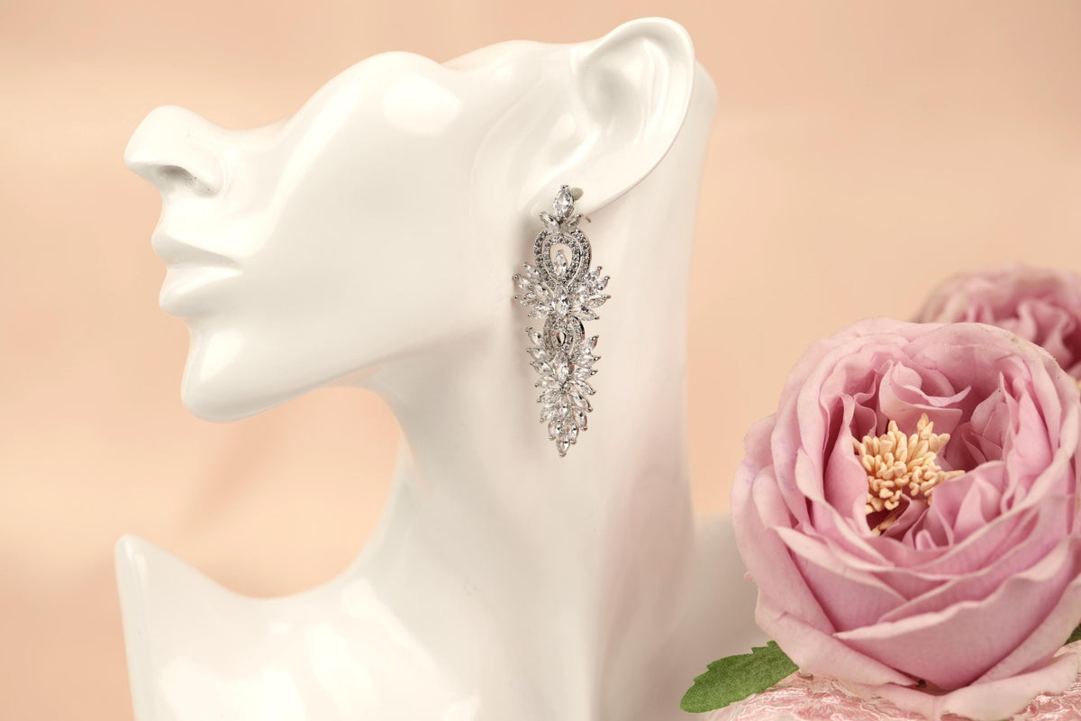 A white mannequin head wearing a pair of silver earrings with a floral design, placed next to a pink rose.