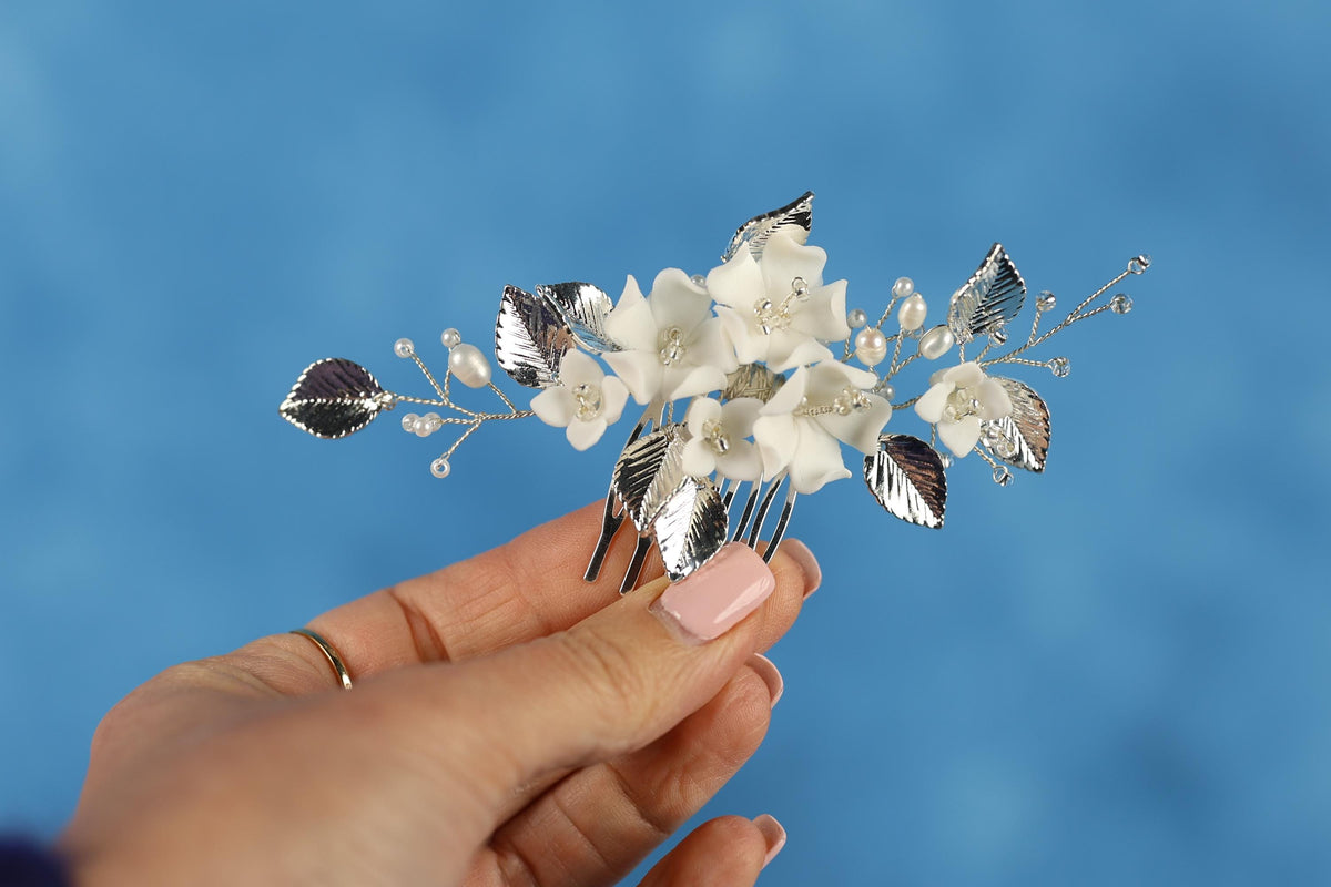 A hand holding a decorative hair clip with white flowers and silver leaves against a blue background.