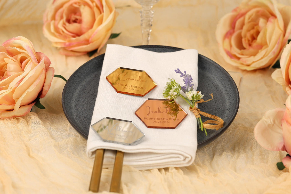a table setting with a white napkin, a black plate, and a glass of wine. There are two hexagonal tags on the napkin, and a small bouquet of flowers is placed on the plate. The table is set against a backdrop of peach-colored roses.