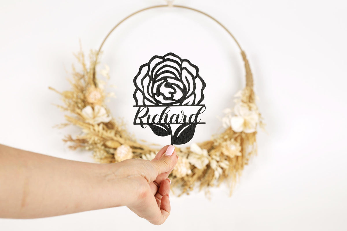 A hand holding a cutout of a rose with the name "Richard" written below it, against a backdrop of a wreath made of dried flowers.