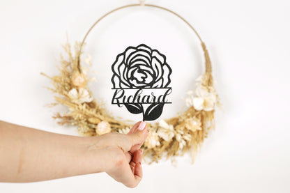 A hand holding a cutout of a rose with the name "Richard" written below it, against a backdrop of a wreath made of dried flowers.