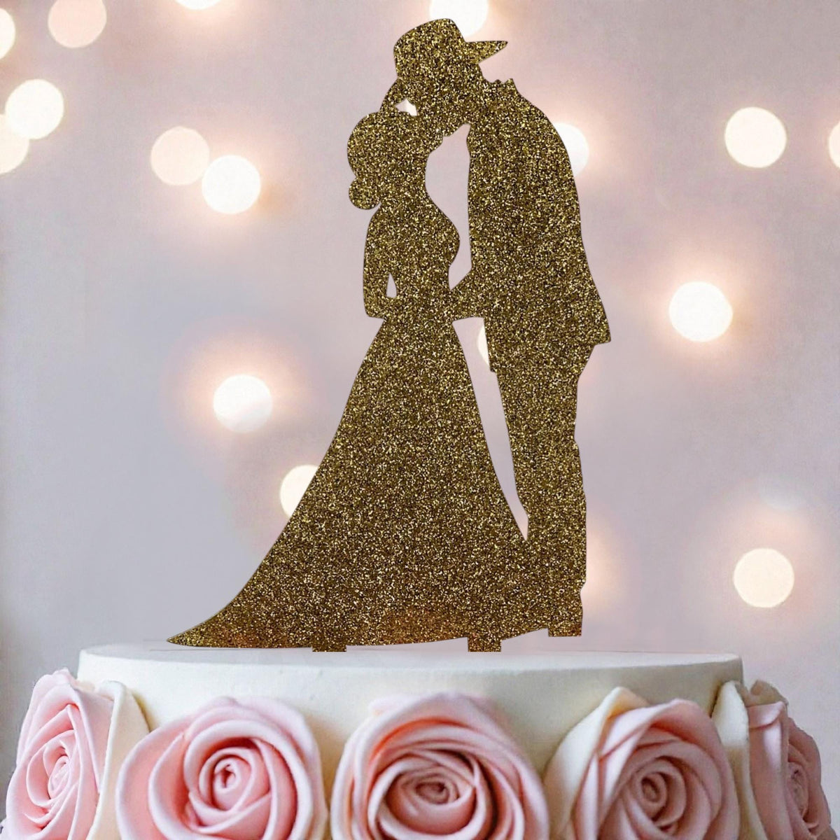 A golden glitter wedding cake topper depicting a bride and groom kissing, placed on top of a white wedding cake with pink roses around the base.