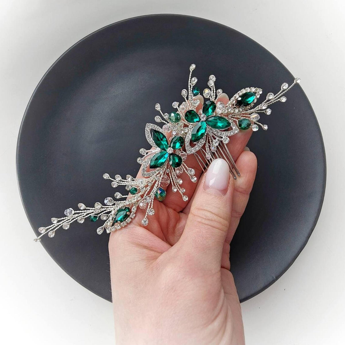 A hand holding a decorative hair accessory with green gemstones on a black plate.