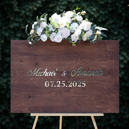 A wooden sign displaying the names "Michael & Amanda" and the date "07.25.2025" against a backdrop of greenery.
