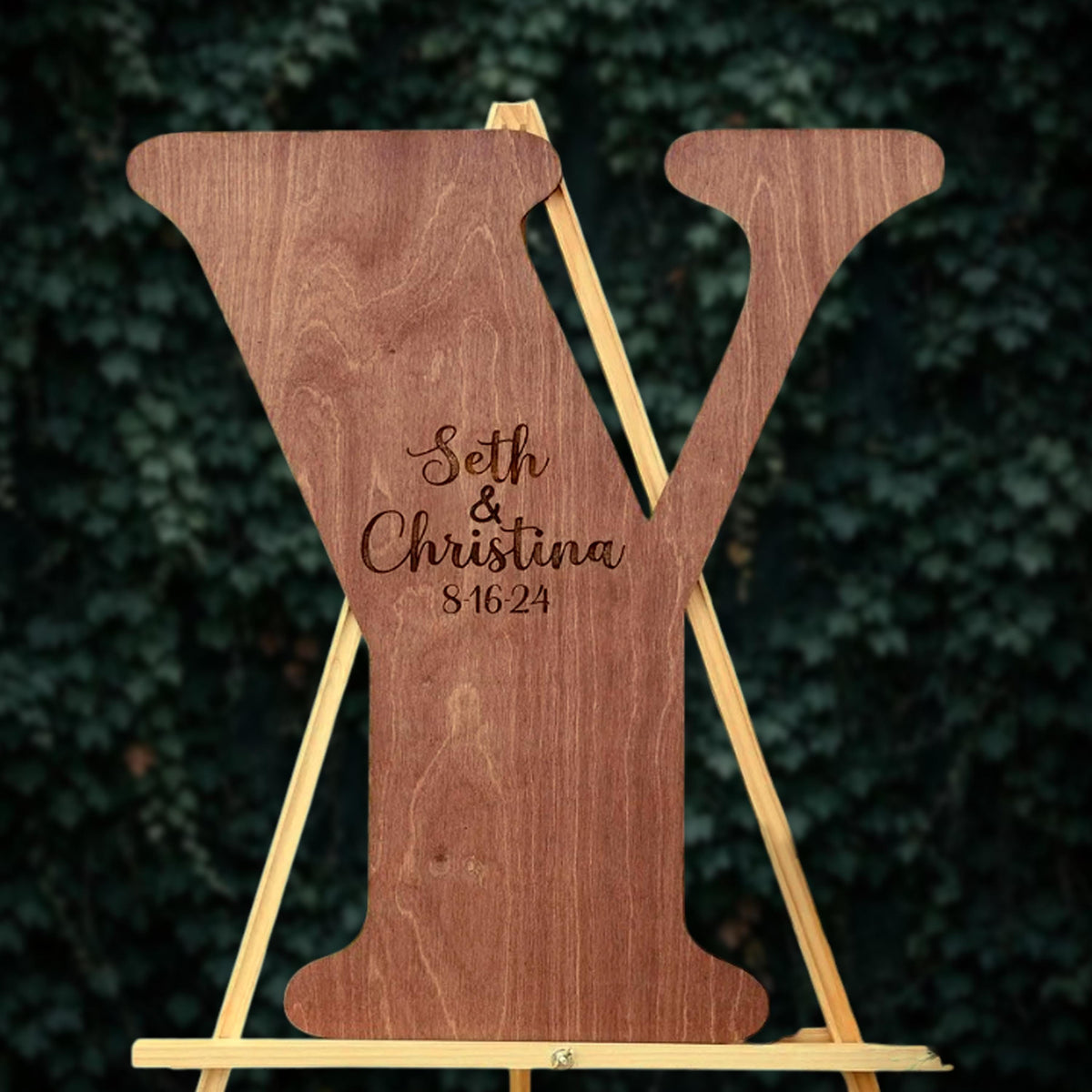 A wooden letter Y with the names Seth and Christina engraved on it, standing on a wooden stand against a background of greenery.