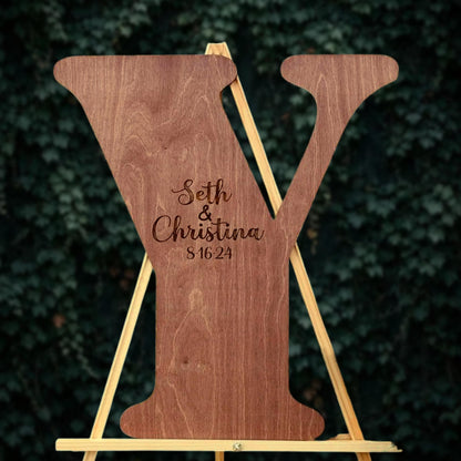 A wooden letter Y with the names Seth and Christina engraved on it, standing on a wooden stand against a background of greenery.