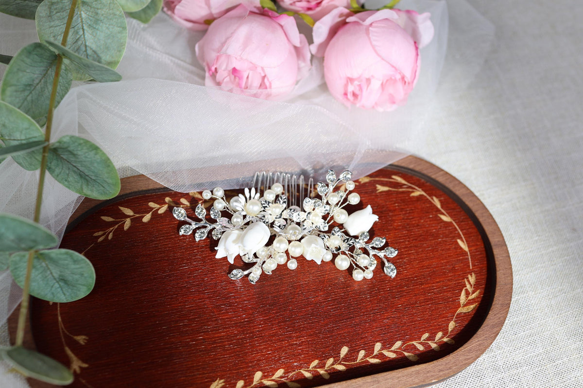 a decorative hair accessory, possibly a hair clip or headband, adorned with white flowers and pearls, placed on a wooden tray. The tray is positioned on a white cloth, and there are pink roses in the background, adding a touch of elegance to the scene.