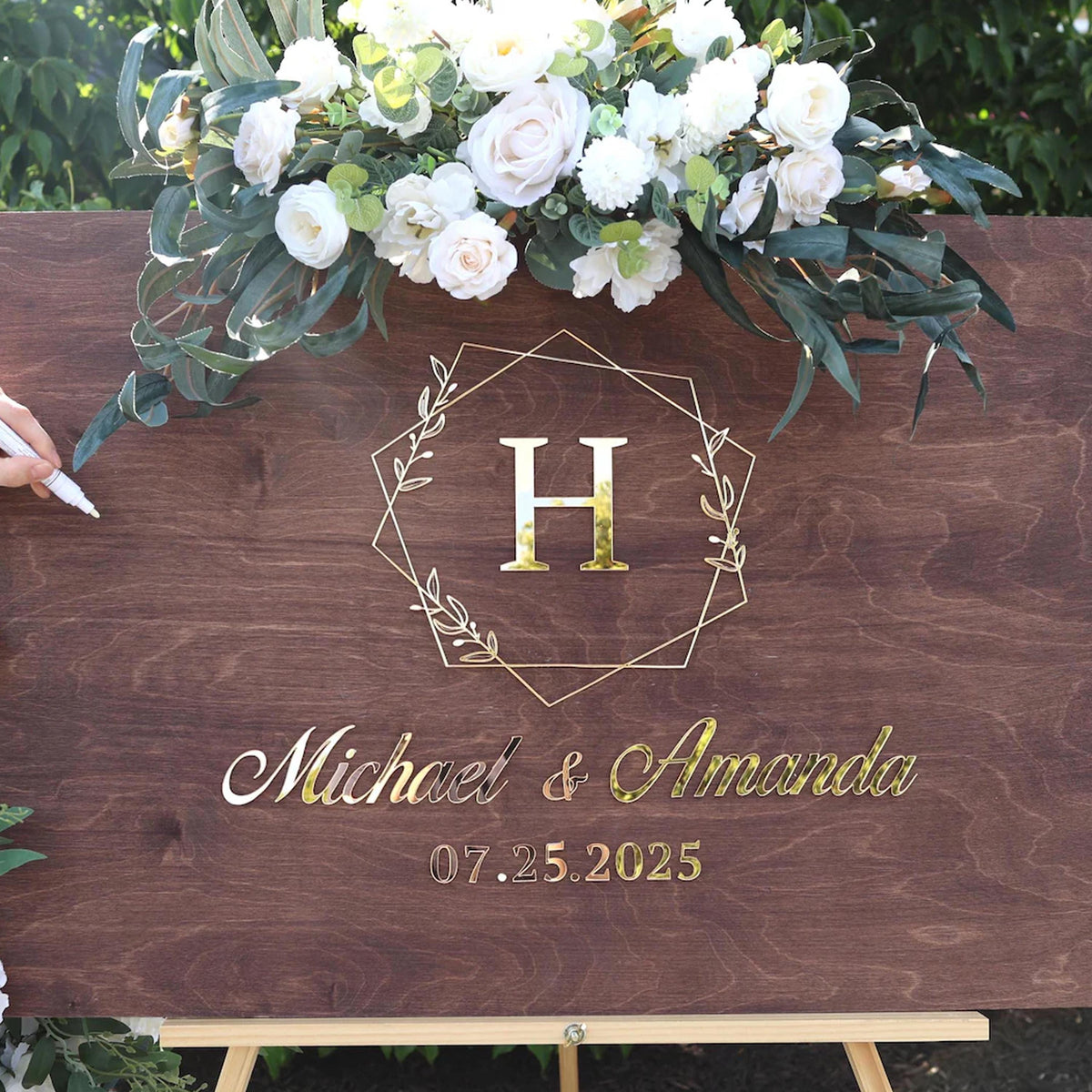 a wooden sign with a floral arrangement and a large letter "H" in the center. The sign displays the names "Michael & Amanda" and the date "07.5.2025".