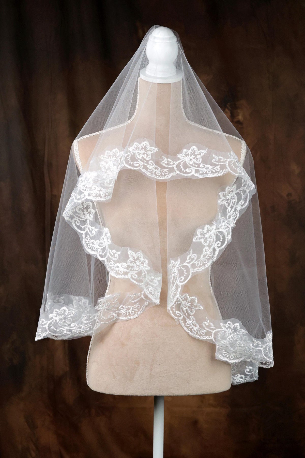 A white wedding veil with intricate lace detailing is displayed on a mannequin against a brown background.