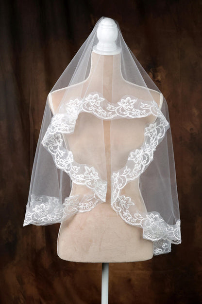 A white wedding veil with intricate lace detailing is displayed on a mannequin against a brown background.