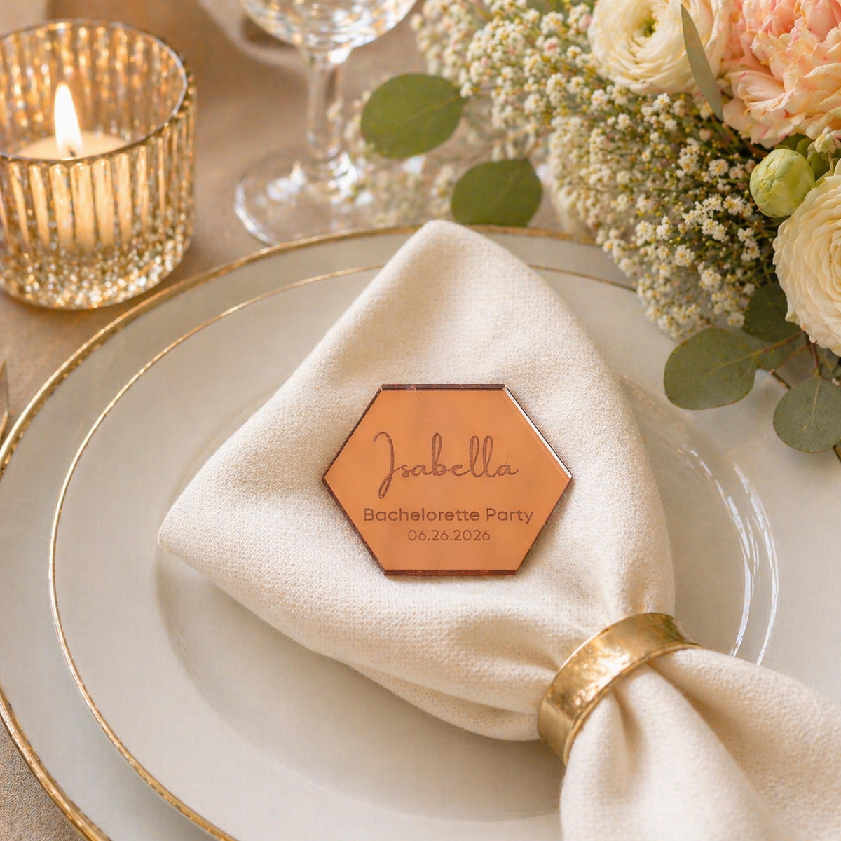 a table setting with a white plate, a gold-rimmed glass, and a candle. There is a hexagonal name tag on the napkin, and a bouquet of flowers in the background.
