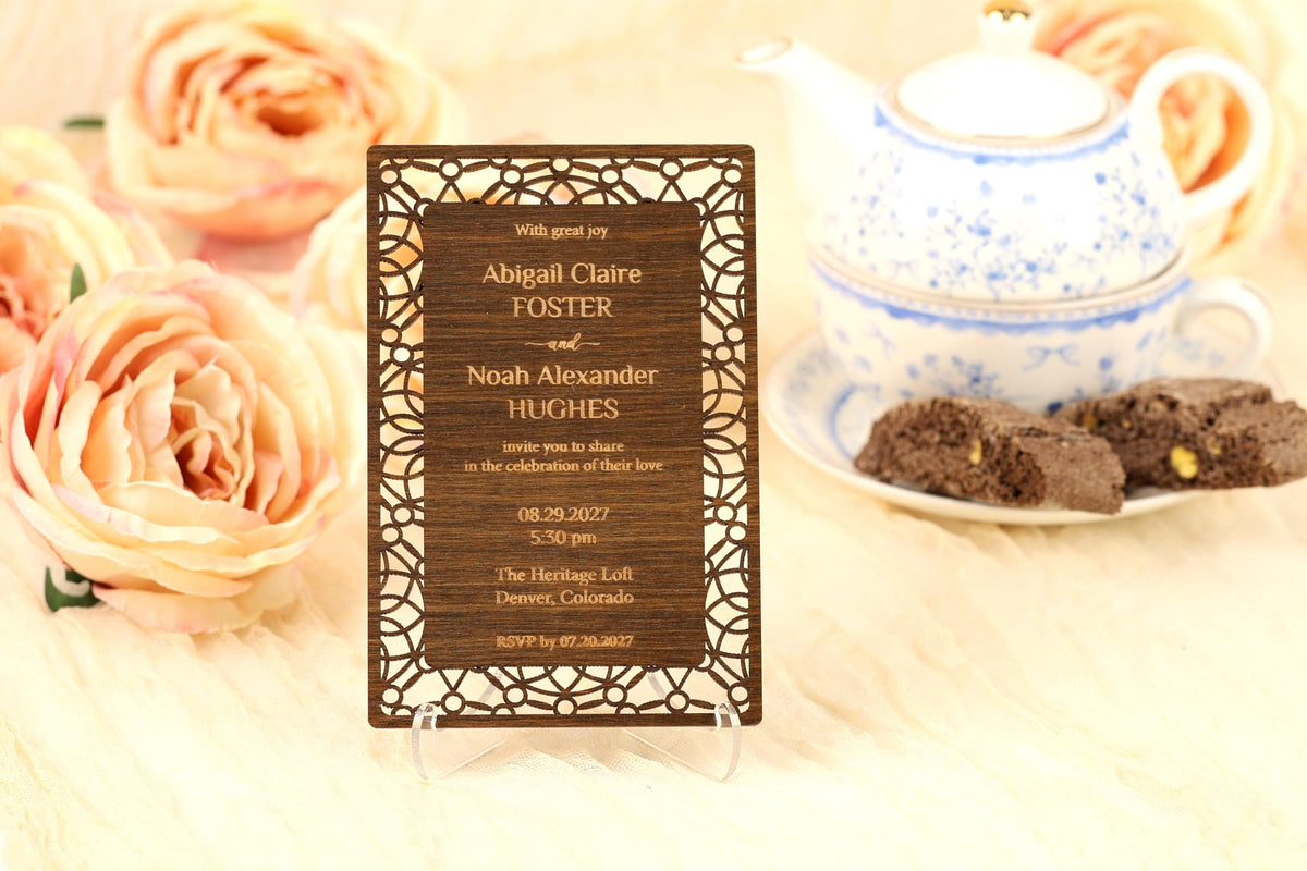 A wooden invitation card with text, placed on a table with a teapot, flowers, and cookies.